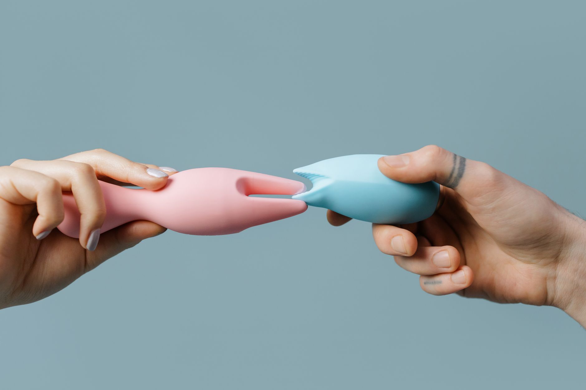 Two hands holding pink and blue silicone vibrators against a pastel background, representing options for clitoral stimulation.